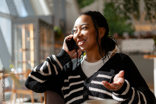 Smiling African American woman holding mobile phone, talking? sitting in cozy cafe, answering call