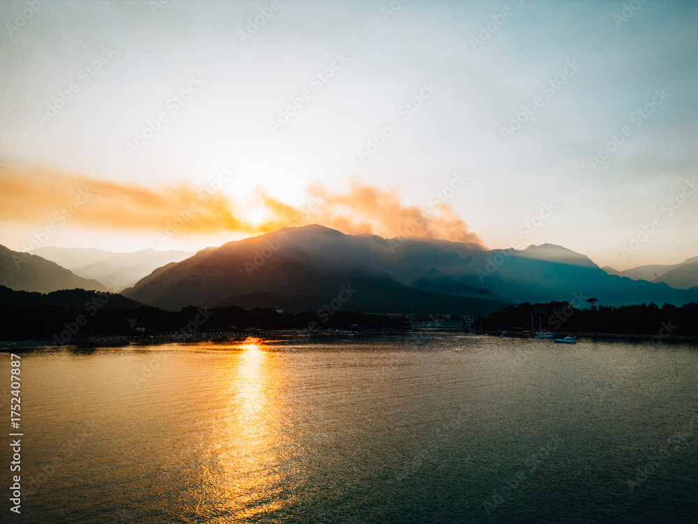 Obraz premium Drone view of sunset with smoke over mountain peaks and golden light reflecting on calm sea. Dramatic landscape with smoky sky, boats, and coastal silhouettes