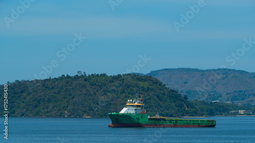 Green and white supply vessel navigates calm waters, with lush hills and mountains forming the backdrop under a clear blue sky