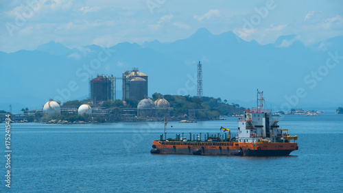 Bunker barge at anchor in calm blue waters near an industrial facility with storage tanks, mountains visible in the distance