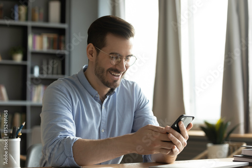 Joyful young male office employee sitting at desk reading phone