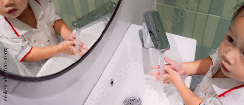 A young child boy wearing pajamas, is actively washing their hands with running water in a modern white sink. In a bathroom, emphasizes hygiene and cleanliness.