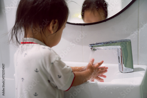 A candid of young child boy in a white shirt and washing their hands under a chrome automatic faucet. The focus is on hand hygiene and the cleanliness routine in a bright, modern bathroom setting.