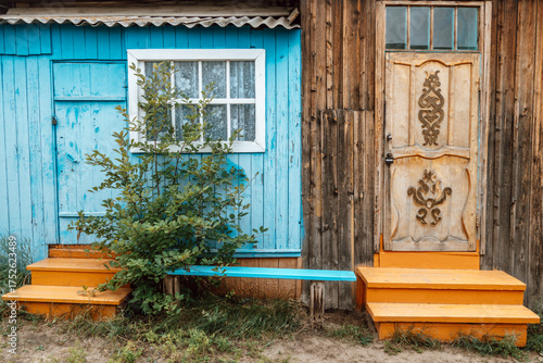 rustic building features bright blue wall with window and an ornate wooden door. orange steps lead to entrance, surrounded by greenery in serene outdoor scene. close up.