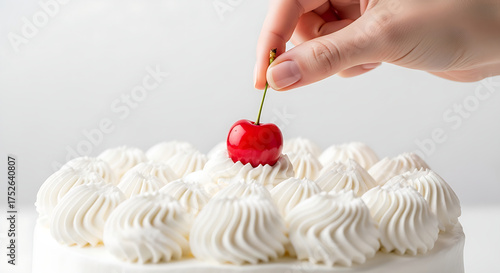 Close up of a hand placing a fresh red cherry on a white frosted cake with swirls of whipped cream isolated on a white background for a decorative dessert concept