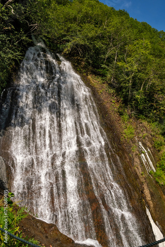 A tall waterfall cascades down a rocky cliff face surrounded by dense summer forest and greenery under a bright sky. Klokovsky Waterfall, Sakhalin Island