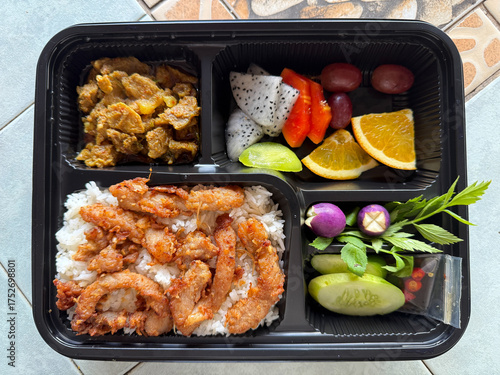 A typical Thai street food lunch box contains jasmine rice, fried pork, pork curry with vegetables, and fruit.