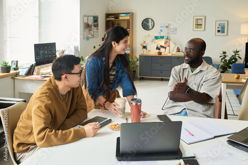 Diverse group of adults including Asian man, woman, Black man collaborating around table in modern office, in discussion with laptops and documents visible