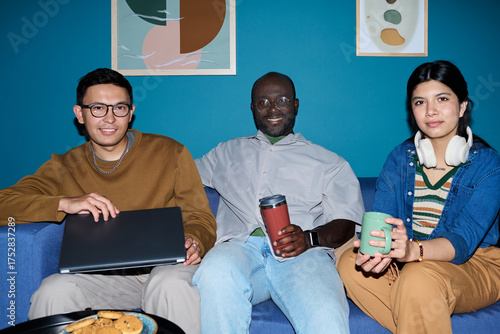 Multiethnic group of adults sitting on couch, smiling and holding coffee mugs and laptop, Black man in center, Asian man on left, woman on right, casual setting