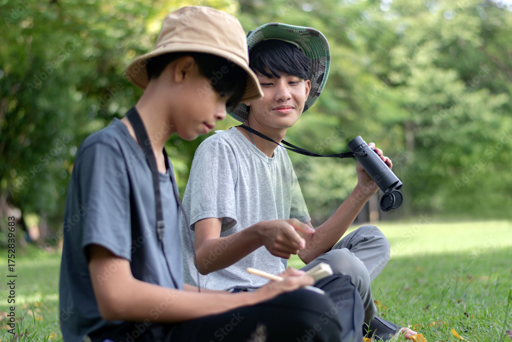 Fototapeta premium Two young explorers collaborate in the field, one observes with binoculars while the other documents the sighting in a notebook.