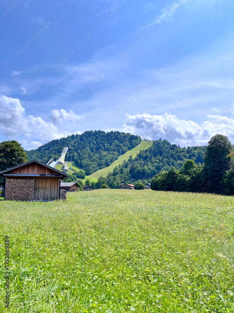 Fototapeta premium Panorama of a wide road that stretches near a mountain settlement through the hills against the background of a sunny blue, slightly cloudy sky.