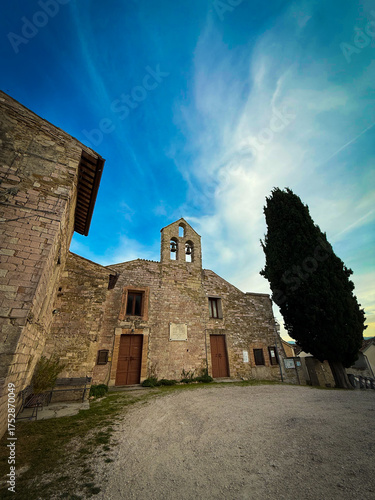A wide-angle, low-view shot of an ancient stone church or monastery with a bell tower, a dirt courtyard, and a tall cypress tree under a vivid blue and cloudy sky.
