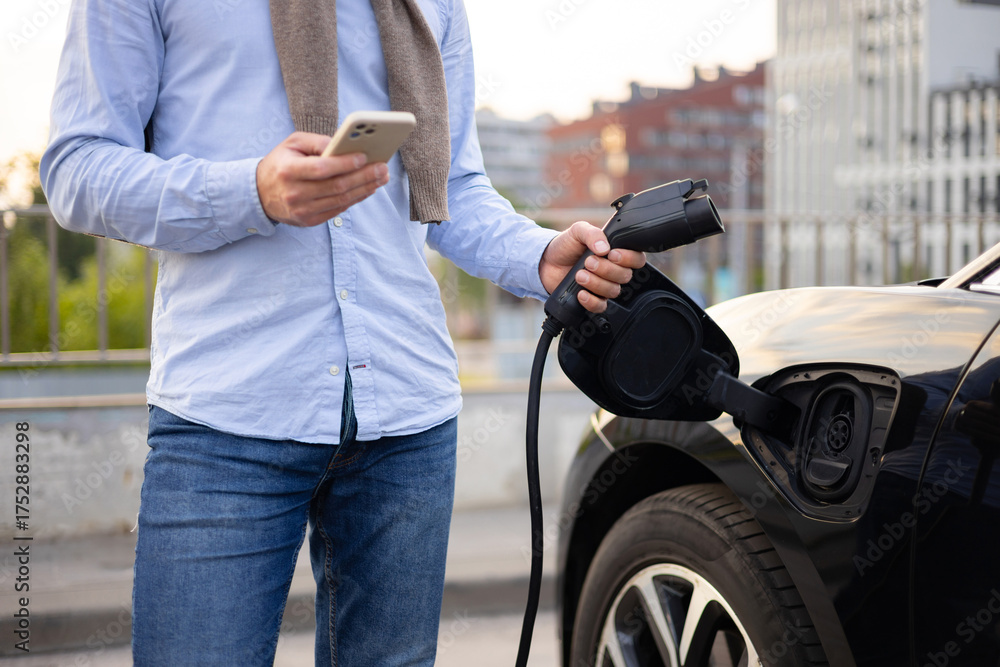 © sofiko14 - A man holding an EV charging cable and using his smartphone near a black electric vehicle. © sofiko14 - A man holding an EV charging cable and using his smartphone near a black electric vehicle.