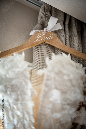 Elegant close-up of wedding dress on wooden hanger with word Bride and white ribbon. Delicate lace fabric and soft light create romantic bridal preparation atmosphere.