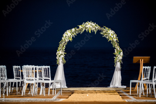 Romantic wedding arch decorated with white flowers and fabric by sea at night. Elegant ceremony setup with chairs and aisle creating beautiful atmosphere for celebration.