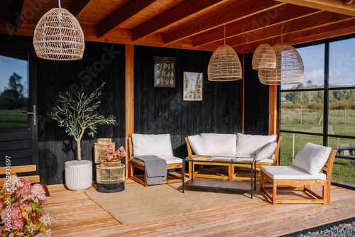 Cozy outdoor seating area with wooden furniture, rattan lights, potted plants, and black wood paneling under a wooden pergola in a peaceful natural setting.