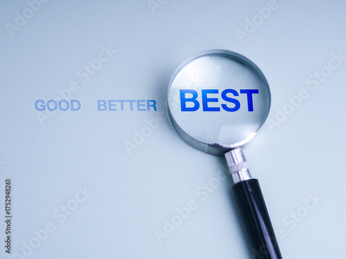 Photography Magnifying glass highlighting the word BEST from a sequence GOOD, BETTER, BEST on blue background, symbolizing excellence, improvement, strategic planning, work development, and decision making
