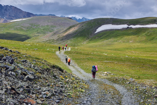 group of hikers makes their way along a winding trail in a lush valley surrounded by mountains. sky is partly cloudy, adding a dramatic backdrop to the scenery. Russia, Altai Mountains