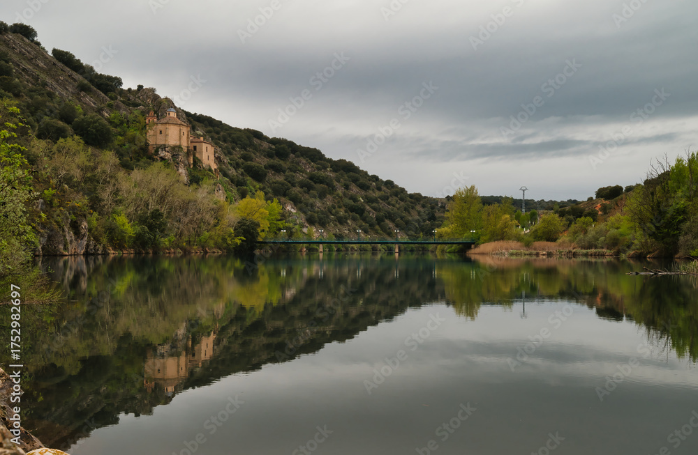 Fototapeta premium The Hermitage of San Saturio is located in a natural setting on the banks of the River Duero, with its reflection in the river, built on a cave on the side of a hill. Soria, Spain. 