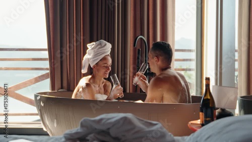 Romantic couple enjoying a hot bubble bath together while clinking champagne glasses in a rustic wooden cabin. Morning light pours in through the window, creating an intimate and serene atmosphere