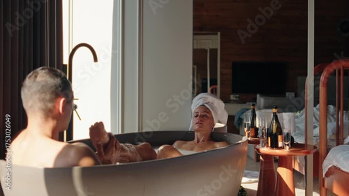 Romantic couple enjoying a hot bubble bath together while clinking champagne glasses in a rustic wooden cabin. Morning light pours in through the window, creating an intimate and serene atmosphere