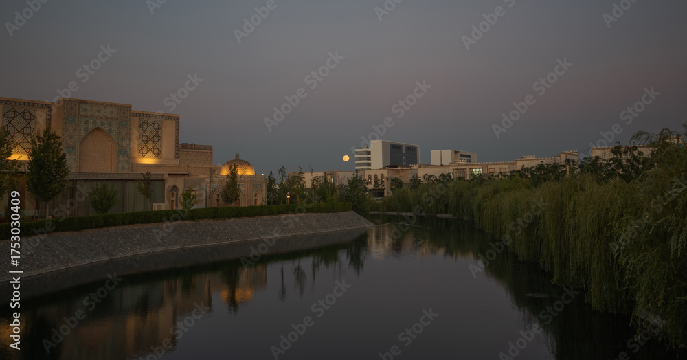 Fototapeta premium Silk Road complex in Samarkand at twilight with illuminated traditional architecture, willow trees along a canal, modern buildings in the distance, and full moon rising above the skyline of Uzbekistan
