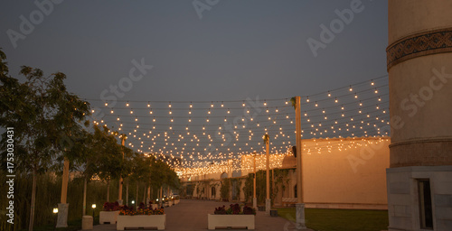 Pathway in the Silk Road complex of Samarkand at dusk with decorative street lamps and trees, illuminated by hanging string lights above, with flower boxes and historic walls in the background.