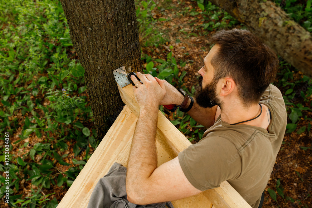 Fototapeta premium brutal young brunette man, dad with beard is nailing building a tree house to his little son with wooden planks, electric red professional drill, self-tapping screws in the forest