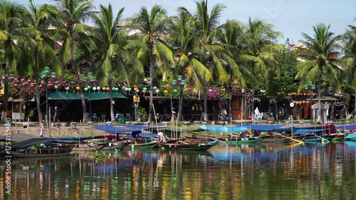 Riverside shops and cafes in Hoi An, Vietnam, feature colorful lanterns and traditional architecture, sheltered by tall palm trees, with tourist boats moored in the foreground.
