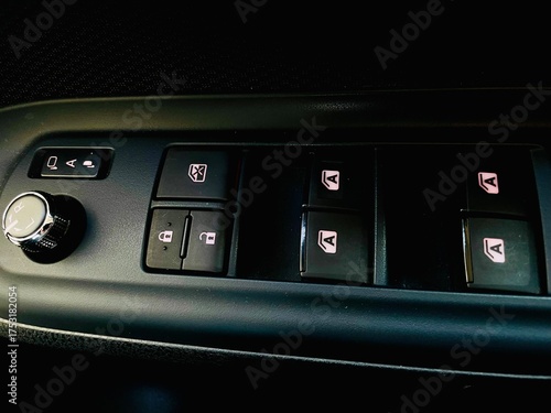 A close-up view of a vehicle's driver-side control panel, featuring power window switches, door lock/unlock buttons, a window lock button, and a knob for adjusting the side mirrors.