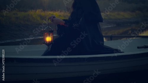 close-up hand holding lantern with warm light, fantasy gothic woman witch floating in boat down river in night autumn nature fog horror dark trees. Mystery girl in ship along lake. Black dress cape
