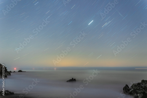 Long-exposure night shot: streaking stars above smooth Atlantic waves and dark Irish coastal rocks.