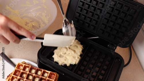 Person puts waffle batter into waffle maker with spatula. Dough spreads inside hot iron before baking. Waffle preparation shows homemade breakfast and cooking process.