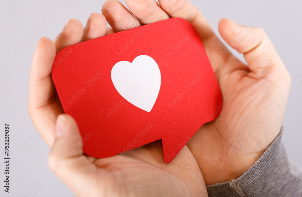 Fototapeta premium Close-up shot of hands gently holding a red heart-shaped speech bubble. Represents love, affection, support, and social interaction. Ideal for V