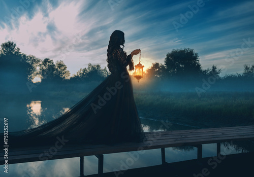 photo real person silhouette gothic woman hand hold lantern lamp walking across bridge over river dense fog mist night nature dark dramatic blue sky lake. long black dress back rear view. Fantasy girl