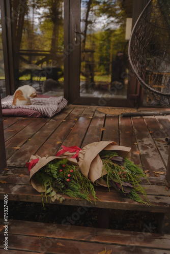 Coniferous bouquets are lying on a wooden terrace near a cozy A-shaped house. Christmas bouquets in kraft paper. Rainy weather. Vertical photo. 