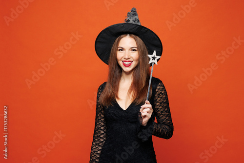 Fotografía Young smiling woman she wear black hat dress witch costume hold magic wand fairy stick look camera isolated on plain red orange background studio portrait
