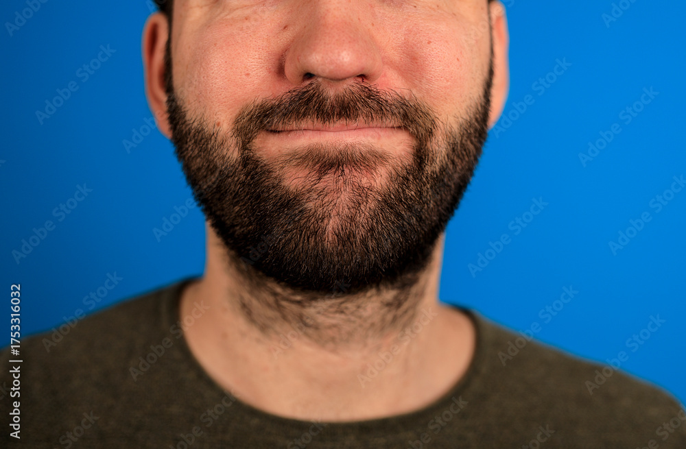 Obraz premium A close-up shot of a man's face with a dark beard, smiling, and a blue backdrop. The image highlights the man's facial features and beard in a s