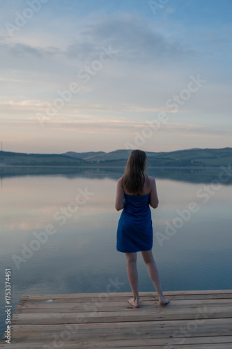 A woman wrapped in a blue towel, on a wooden dock by a calm lake during sunset.
