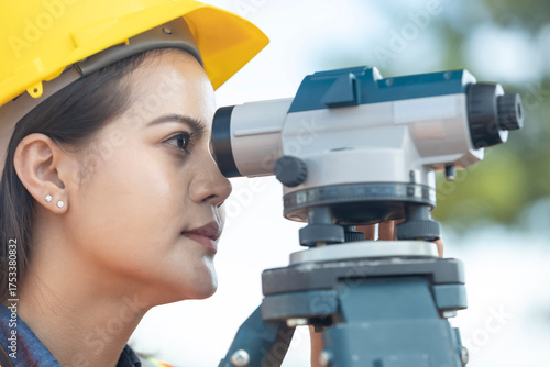 Close-up of beautiful Asian woman wearing a safety helmet using surveying equipment at construction site. Side view.