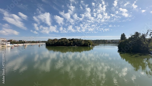 Fényképezés Calm river scene in Vinnytsia, Ukraine, with a small island, boats, and urban shoreline under a blue sky with altocumulus clouds