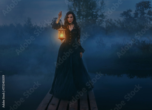 Fairy tale mystery woman queen hand hold lantern lamp candle walking across bridge pier over river dense fog mist dark blue night nature. Fantasy girl beauty face gothic makeup long hair black dress