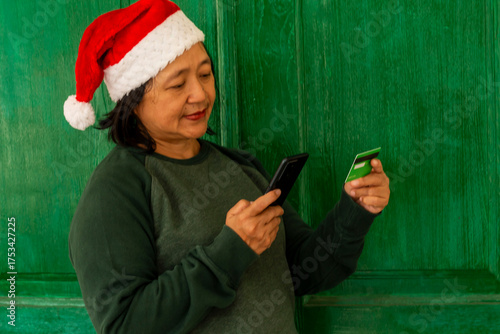 An adult woman, wearing a green sweater and a red Santa Claus hat, is making a transaction using a mobile phone. A green credit card. A green background.
