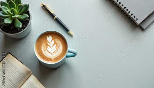 Modern desk setup with light blue coffee mug showing latte art. Green succulent plant, pen, open book, spiral notebook placed on clean table. Calm workspace great for focused study, work, relaxing at