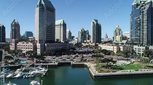 Smooth aerial drone tracking video, high angle, sweeping over vibrant San Diego waterfront. Majestic glass skyscrapers reflect bright blue sky, bustling marina with white boats, people on bridge.