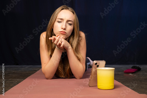 A young woman lies on a yoga mat with a quizzical look, engaging with her phone in an expressive moment.