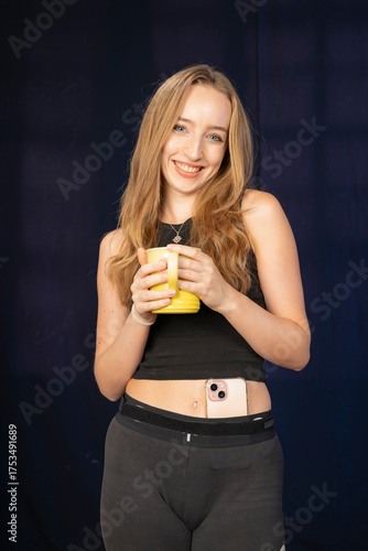 This cheerful young woman has a bright smile while holding a yellow mug and is dressed in a fitted black top and leggings, with her smartphone visibly tucked into her waistband.