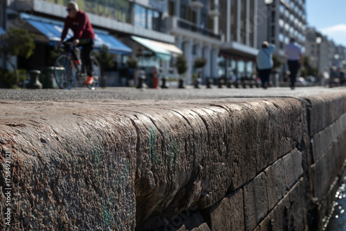 close-up of old stone pier wall in a sunny city, with a blurred background showing a cyclist, pedestrians, and waterfront buildings. urban outdoor scene.