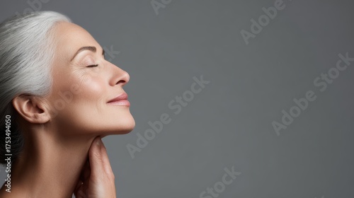 Profile portrait of a serene elderly woman with gray hair, gently touching her chin and smiling with closed eyes. Spa rejuvenation and skincare concept, gray background