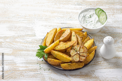 Golden baked potato wedges with garlic, spices and Greek tzatziki sauceon rustic plate. Tasty homemade snack or side dish, healthy vegetarian food